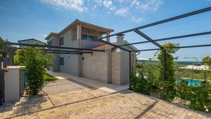 Ein modernes Haus mit Backsteinfassade, einer überdachten Terrasse und Blick auf einen Swimmingpool sowie Hügel im Hintergrund.