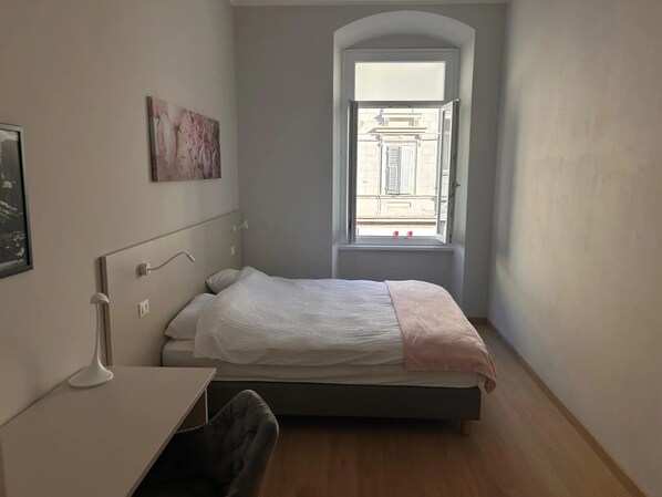 Un dormitorio con cama, escritorio, silla, ventana con arco y lámpara de pared.