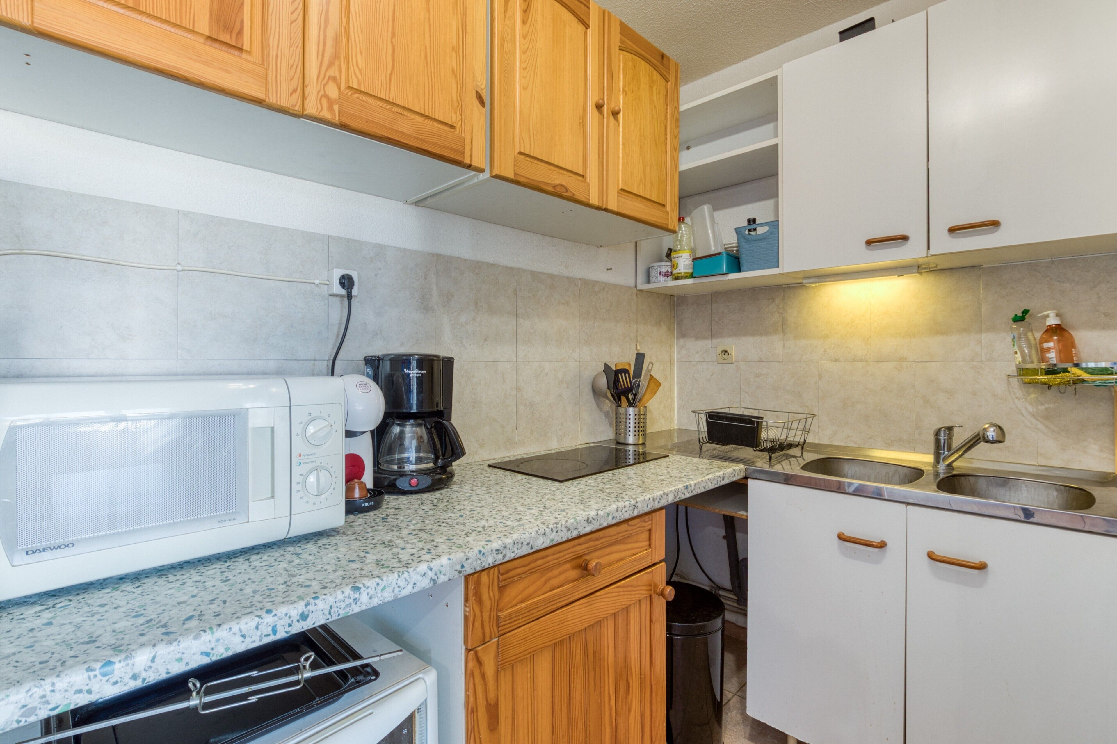 Una cocina con armarios de madera, microondas, cafetera, lavamanos y cubo de basura.