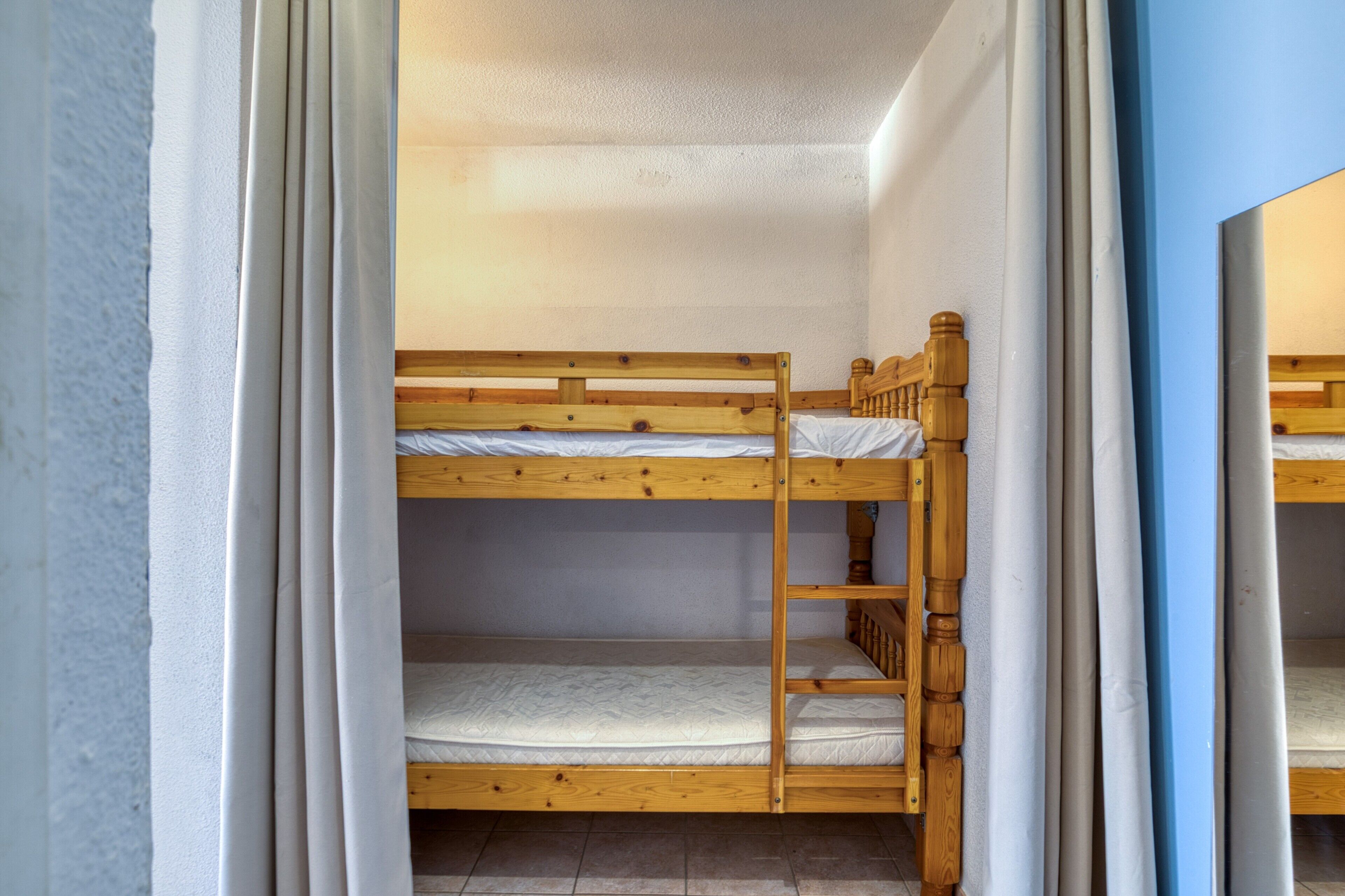 Dormitorio con literas de madera, cortinas blancas y suelo de baldosas.