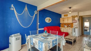 A dining area with a table set for two, a red chair, a hammock, and a kitchen area in the background. - “L’Escale Marine” – AC, Beach, Sleeps 4 (Sète)