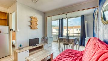 Een kamer met een rode bank, een houten tv-meubel, een eettafel met stoelen en een balkon met uitzicht op gebouwen.