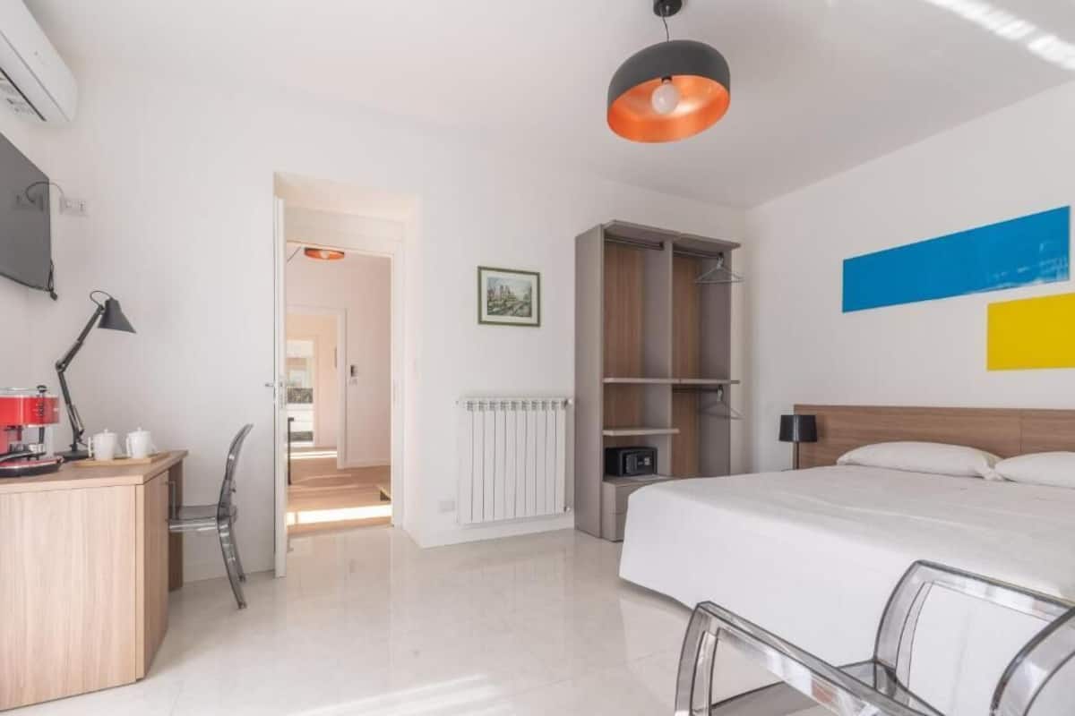 A modern hotel room with a bed, a desk, a chair, a radiator, and a wardrobe.