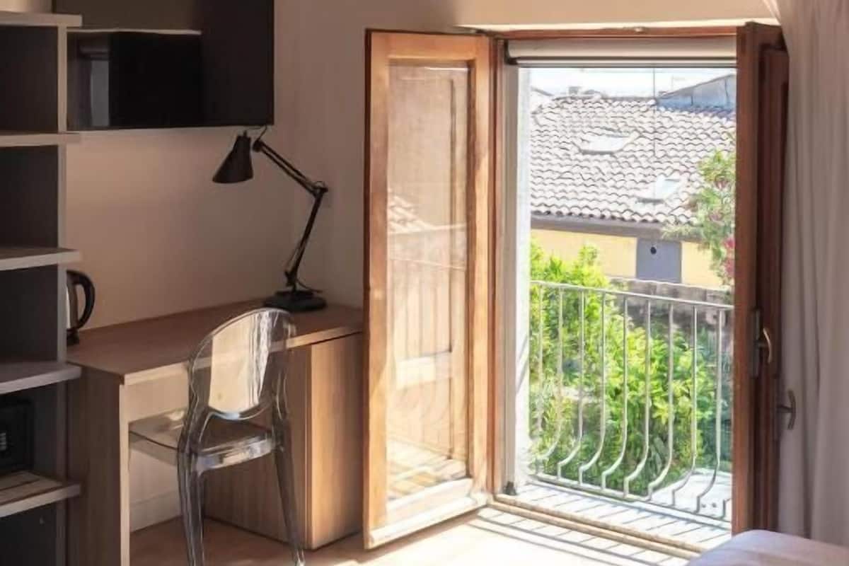 A bedroom with a wooden ceiling, a bed, a desk with a chair, a TV, and a balcony with a view.