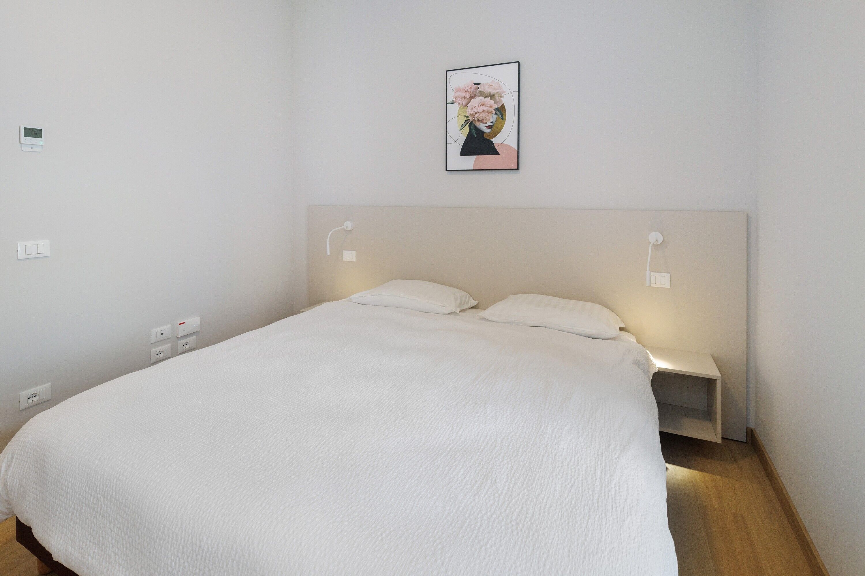 Een net opgemaakt bed met witte lakens, een hoofdbord, nachtlampjes en een wandcontactdoos.
