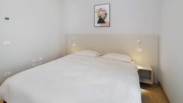 Een net opgemaakt bed met witte lakens, een hoofdbord, nachtlampjes en een wandcontactdoos.