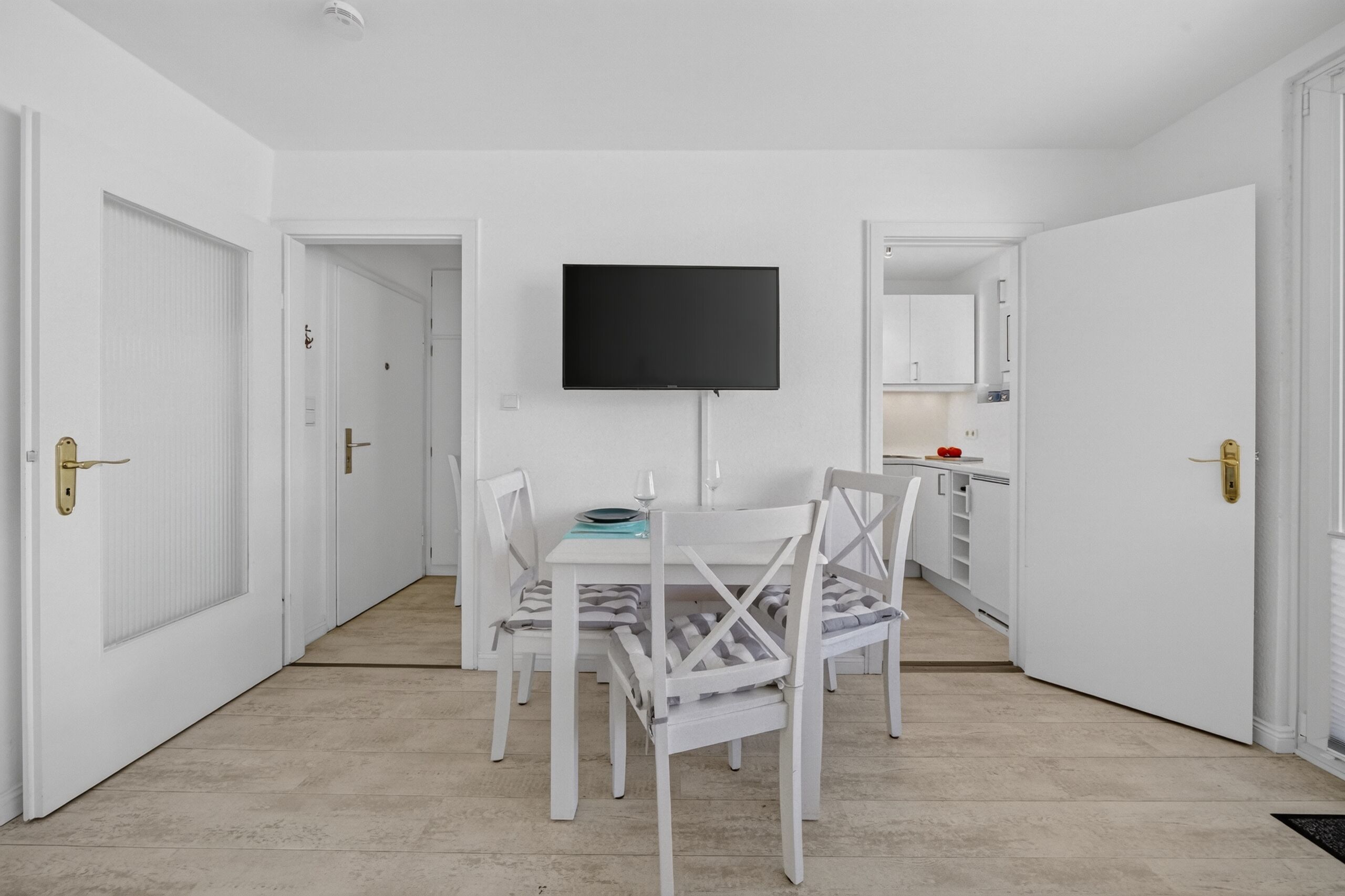 A room with a dining table, chairs, a flat-screen TV mounted on the wall, and a kitchen area visible through an open door.