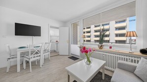 A modern, well-lit room with a flat-screen TV, white furniture, a dining area, a large window with a view of buildings, and a radiator. - 2-Bedroom Vacation Apartment Near the Beach in Westerland (Sylt)