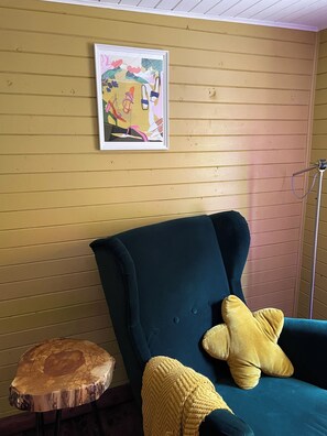 Una stanza con un divano color petrolio, una coperta gialla lavorata a maglia, un quadro astratto incorniciato e uno sgabello di legno.