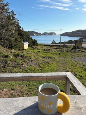 Un banco de madera con una taza, con vistas a un paisaje costero con árboles y un cuerpo de agua.