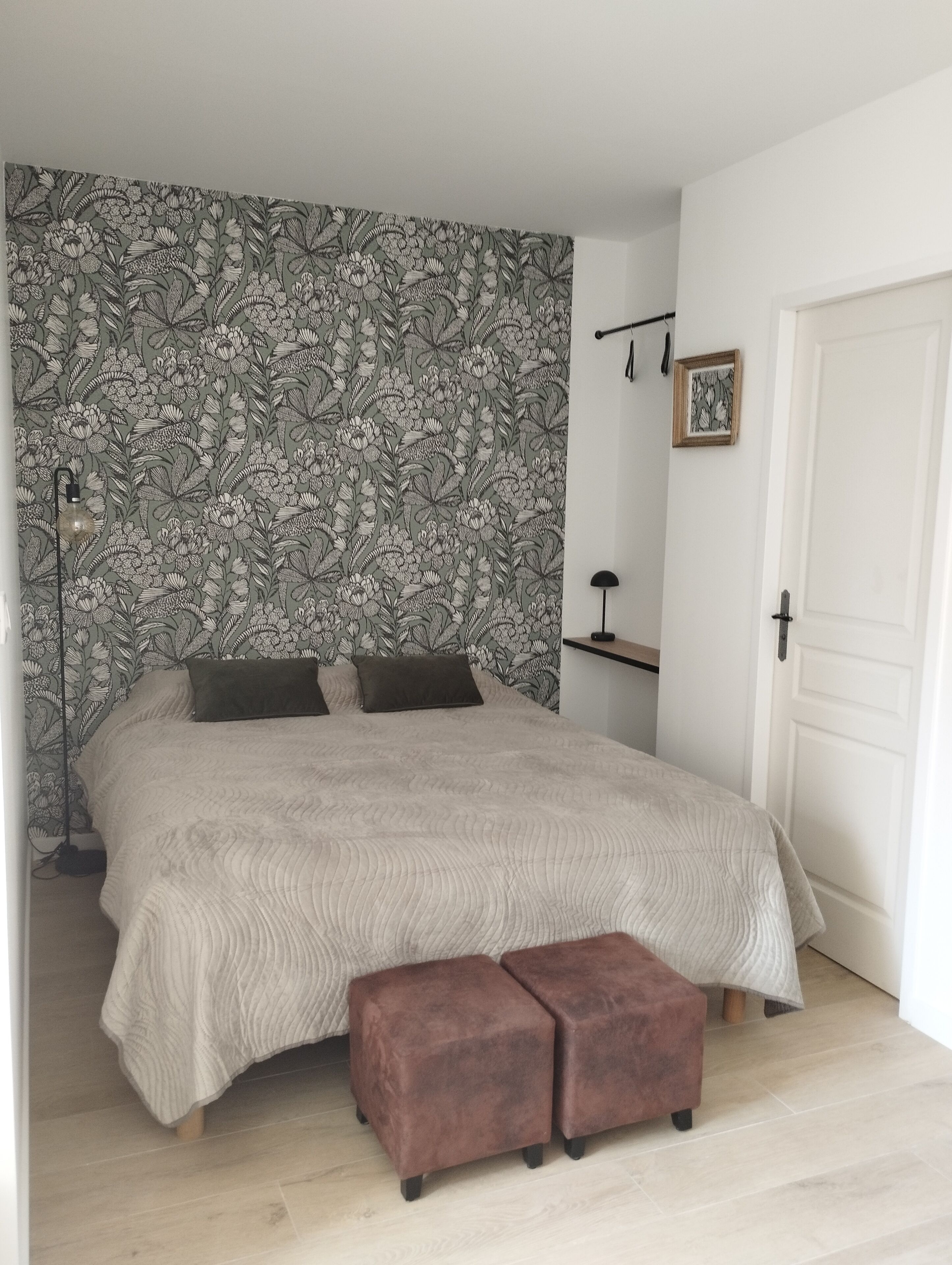 A bedroom with a floral wallpaper, a bed with a beige quilt, two brown ottomans, a bedside lamp, and a white door.