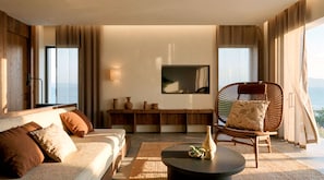A modern living room with a sofa, a round coffee table, and a wicker chair. There is a flat-screen TV mounted on the wall, and a window with curtains. - Ella Elyra - Adults Only - All Inclusive (Corfu)