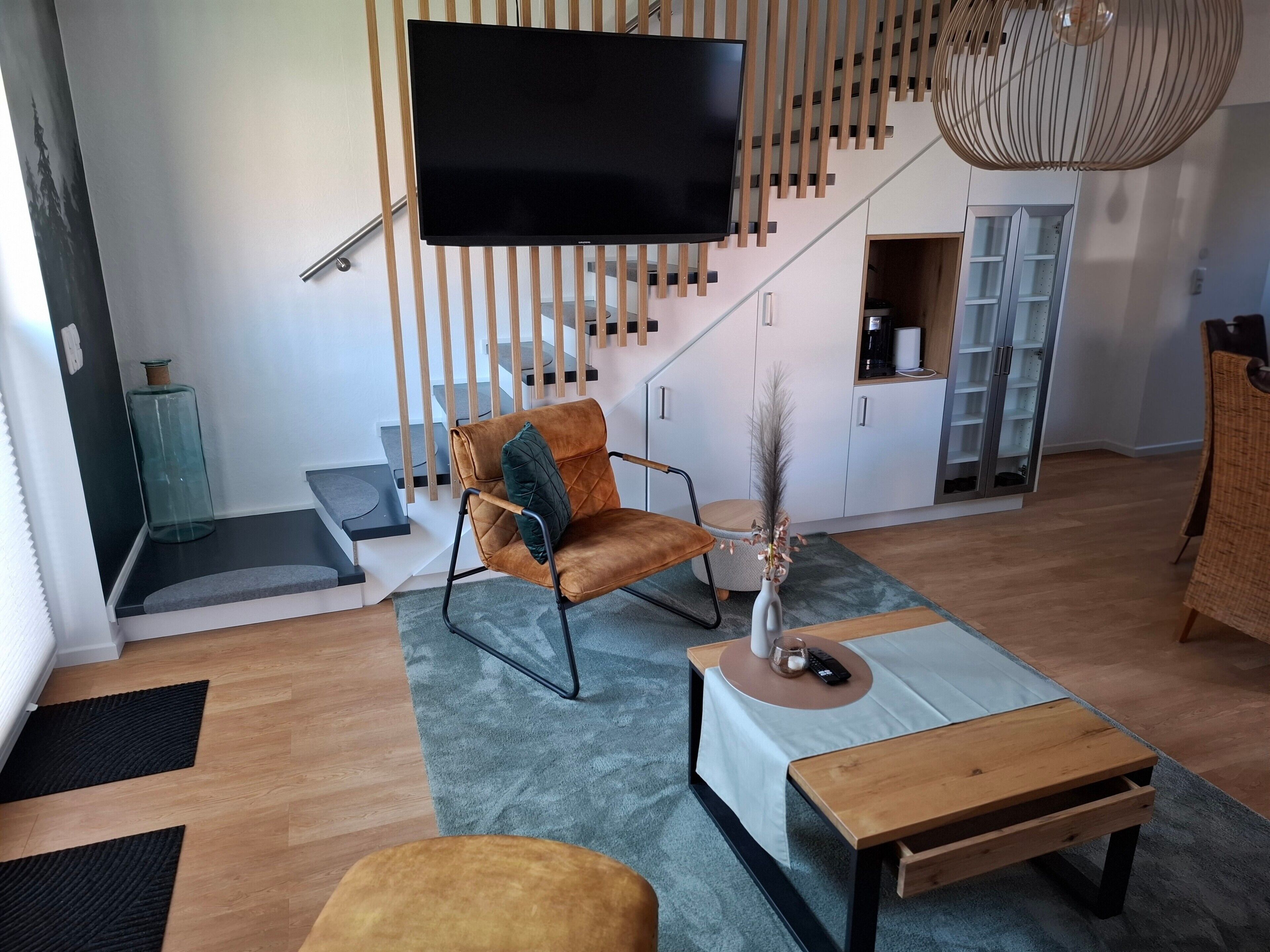 Een moderne woonkamer met een flatscreen televisie, een bruine leren fauteuil, een houten salontafel en een trap met houten leuningen.