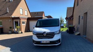 Ein weißer Renault Transporter steht auf einem gepflasterten Einfahrt vor einem Wohngebiet mit Ziegelfassaden und blauem Himmel.