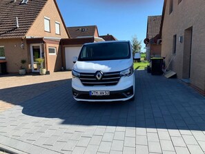 Ein weißer Renault Transporter steht auf einem gepflasterten Einfahrt vor einem Wohngebiet mit Ziegelfassaden und blauem Himmel.