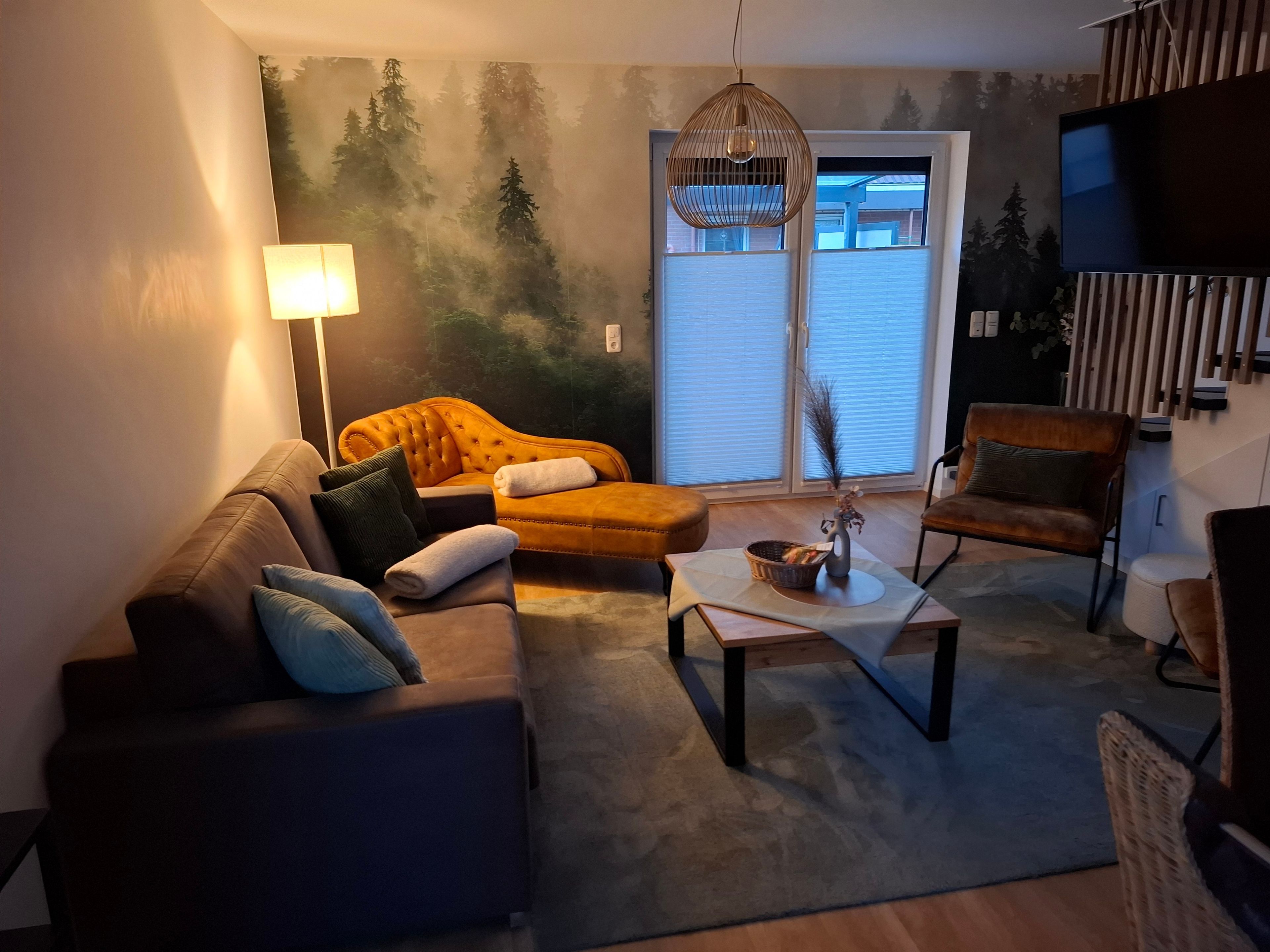 Una sala de estar con sofá, sillón, reposapiés, una mesa de centro, una lámpara y un mural en la pared.