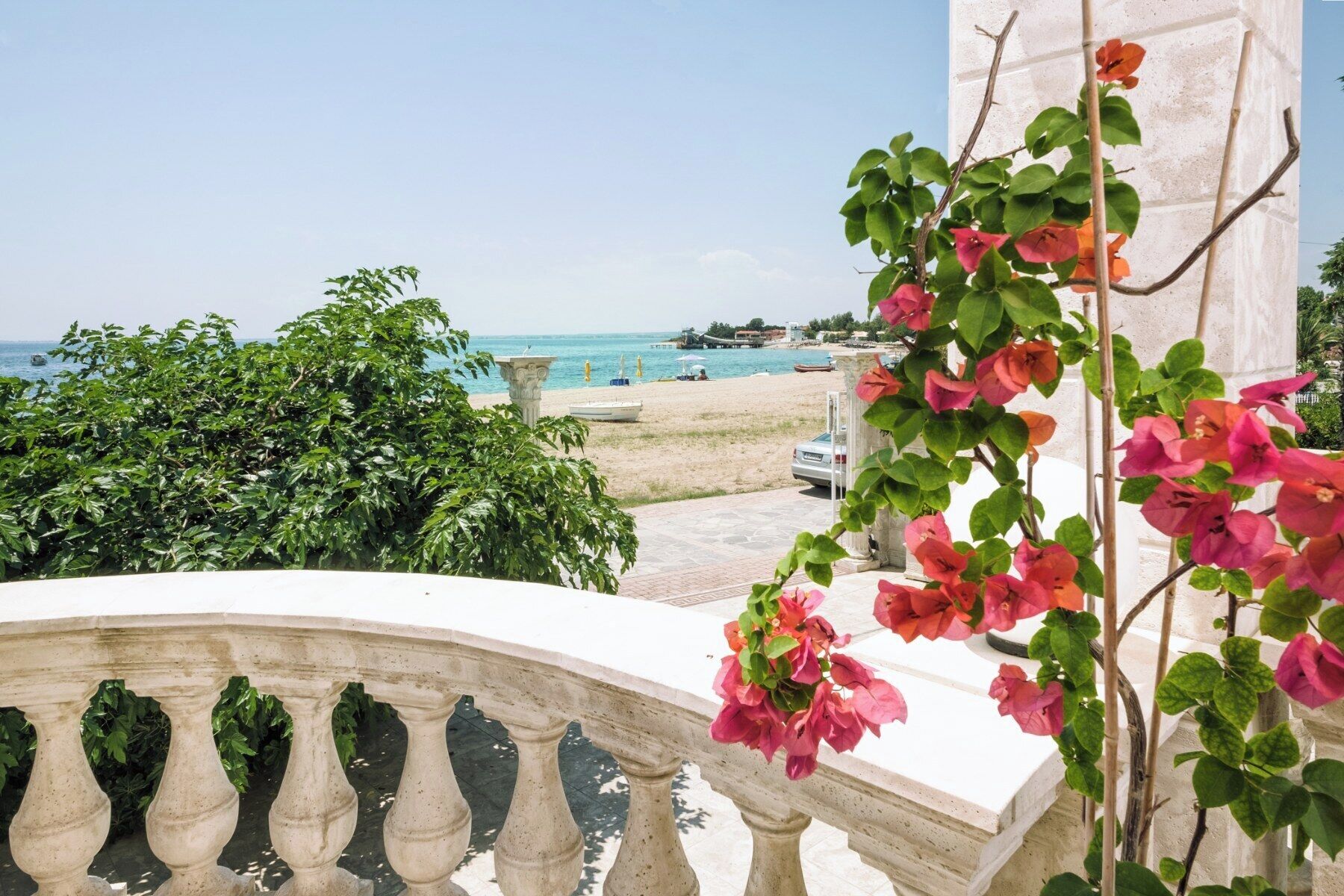 Un balcon avec vue sur la mer, une voiture blanche et des fleurs de bougainvillier.