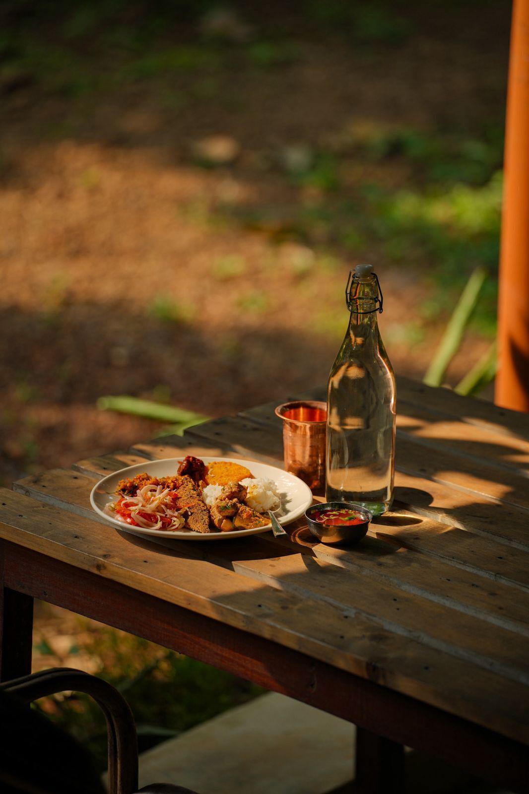 Una mesa de picnic de madera con un plato de comida, una botella y una taza metálica pequeña.