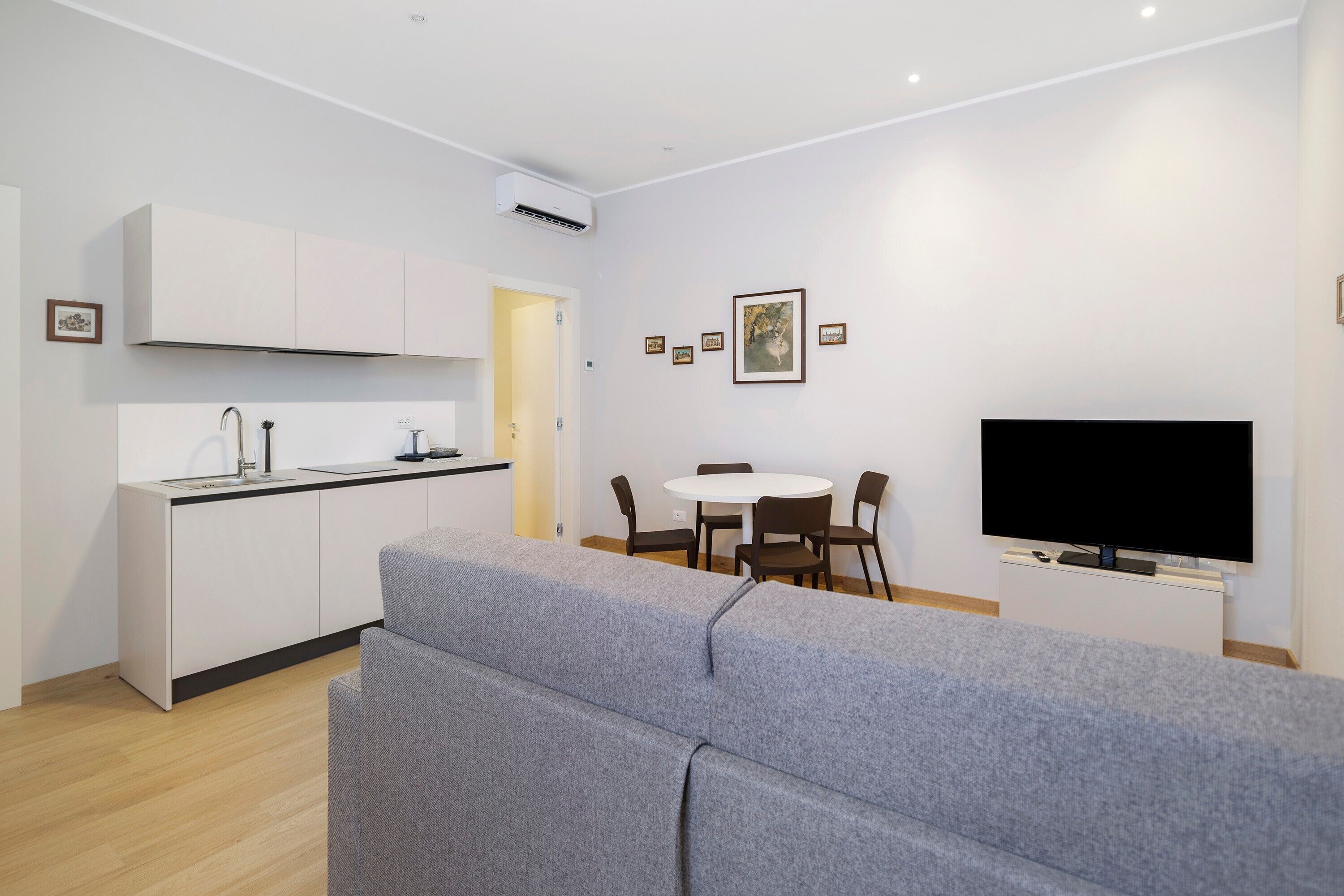 Ein modernes Wohnzimmer mit grauem Sofa, einem Flachbildfernseher, einem Essbereich mit weißem Tisch und Stühlen sowie einer Küche mit weißen Schränken und einer schwarzen Arbeitsplatte.