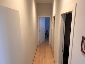 Een smalle gang met houten vloer en witte wanden, die leidt naar een kamer met een deur aan de rechterkant.