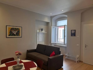 Een woonkamer met een bank, een eettafel met stoelen, een vaas met bloemen, een schilderij aan de muur en een raam met uitzicht.