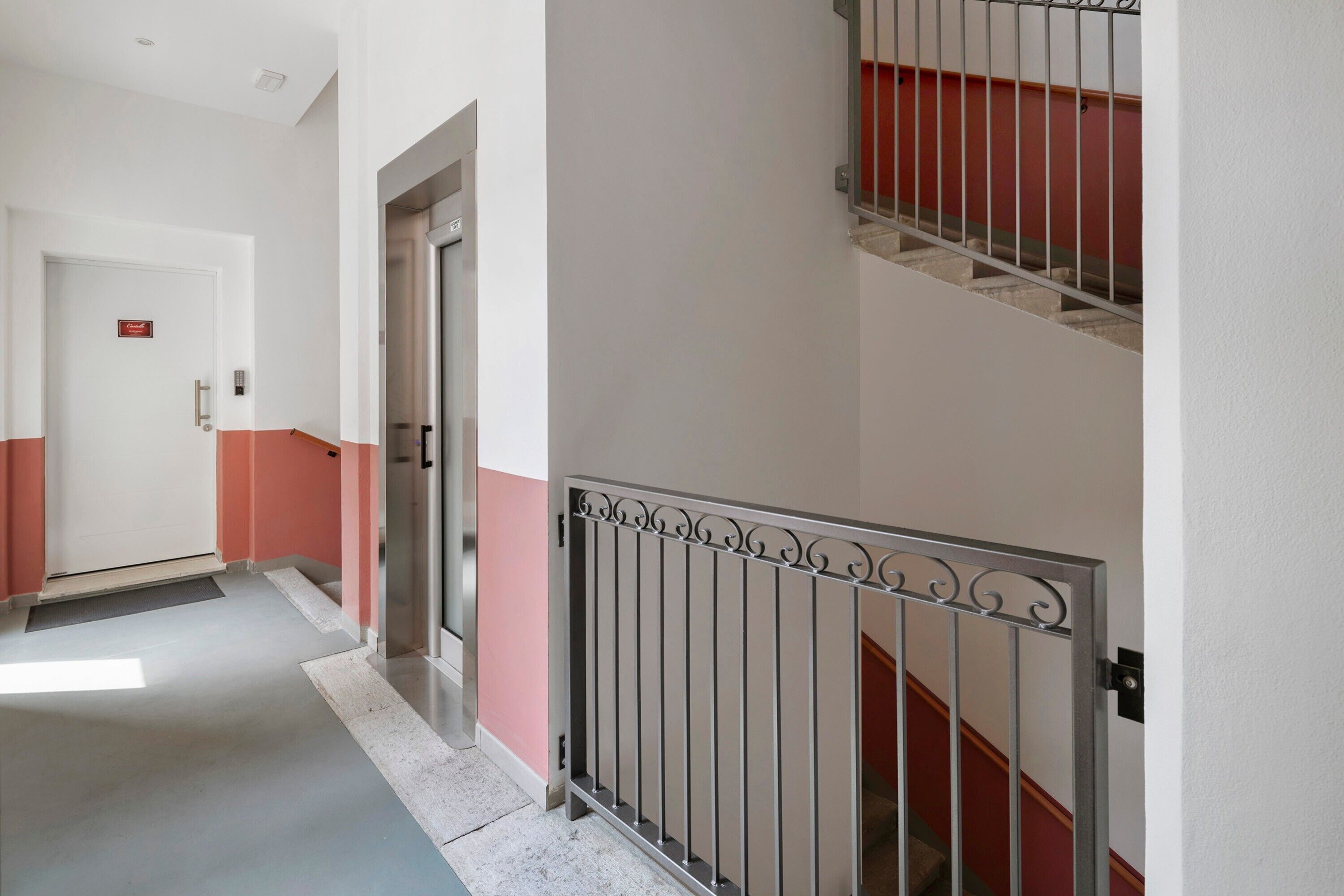 A corridor with a metal railing, a door with a sign, and a staircase with a red handrail.