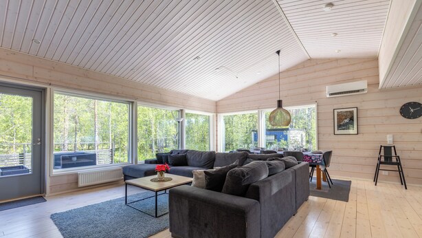 Un salon moderne avec un grand canapé d’angle, une table basse et un coin repas comprenant une table et des chaises. De grandes fenêtres laissent entrer la lumière naturelle et offrent une vue sur l’extérieur.