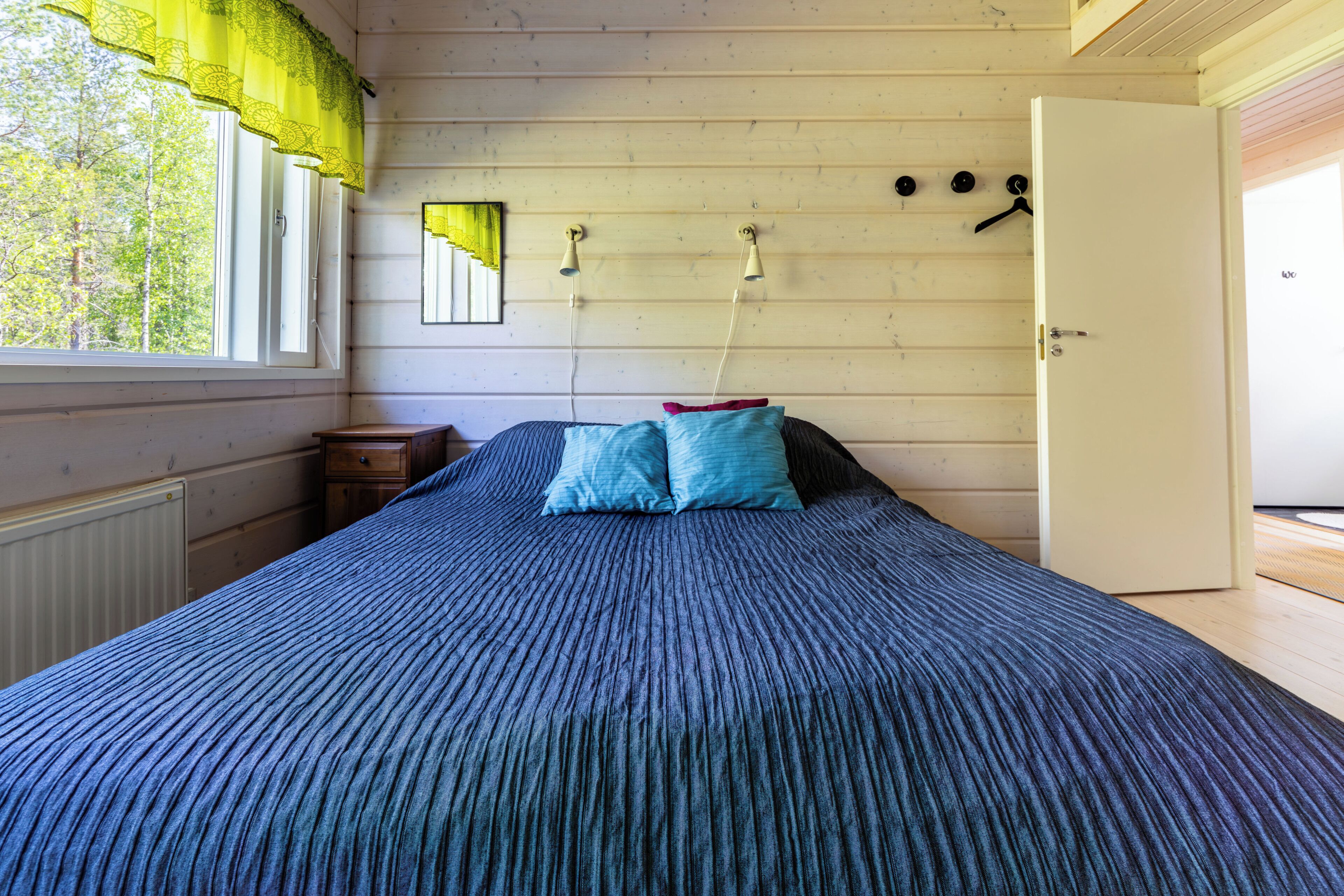 A bedroom with a bed, a window with curtains, a radiator, a door, and a wall-mounted light fixture.