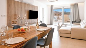 A modern living room with a wooden dining table, grey chairs, a beige sofa, a flat-screen TV, and a balcony with a view of the city. - Modern Seaside Apartments (Zebbug)