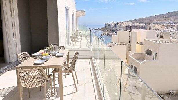 A balcony with a table set for dining, overlooking a coastal cityscape with buildings and boats. - Modern Seaside Apartments (Zebbug)