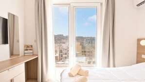 A hotel room with a bed, a desk, a flat-screen TV, and a view of the cityscape through large windows. - Modern Seaside Apartments (Zebbug)