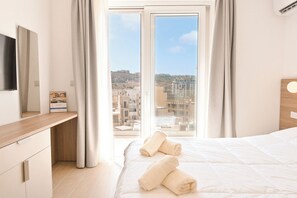 Ein Hotelzimmer mit Bett, Schreibtisch, Flachbildfernseher und Blick auf die Stadt durch große Fenster.
