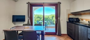 Ein Zimmer mit Esstisch, Stühlen, Fernseher, Küchenzeile und einer Glastür, die auf einen Balkon mit Blick auf Grünflächen führt.