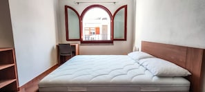 Ein Einzelbett mit Holzvorhang, ein kleiner Schreibtisch mit Stuhl und ein Fenster mit geöffnetem Klappladen.
