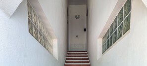 Eine schmale Treppe führt zu einer Tür, über der eine Leuchte angebracht ist, gesäumt von schlichten Wänden und einem Fenster mit Gittermuster.