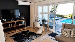 A modern living room with a flat-screen TV, wooden shelves, a glass table, a sofa, and a view of a swimming pool. - Villa Atlantica  (Cape Town)