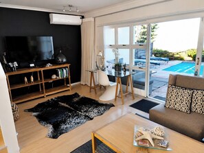 Een moderne woonkamer met een flatscreen televisie, houten planken, een glazen tafel, een bank en uitzicht op een zwembad.