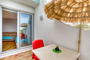 Un balcon avec une table, une chaise rouge, un pot de fleurs et un parasol en paille.