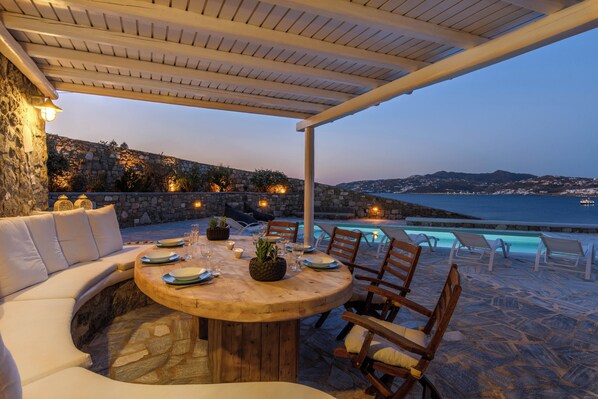 A covered outdoor dining area with stone walls, a pool, and a view of the sea. - Lunar Villa (Mykonos)