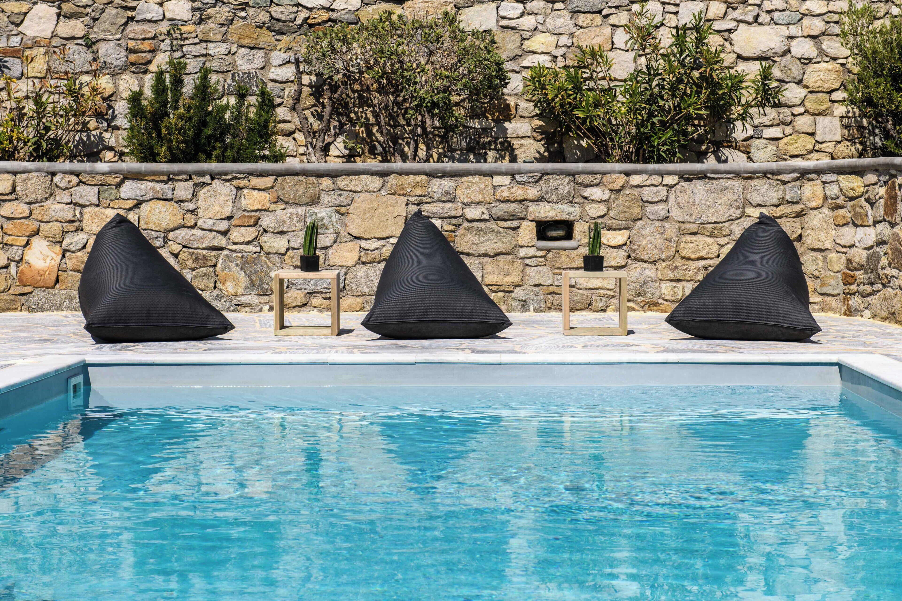 Ein Pool mit drei stuhlähnlichen schwarzen Liegen und einer Steinwand mit Pflanzen.