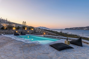 A stone pool with lounge chairs and cushions, overlooking a coastal landscape with a view of the sea and distant hills. - Lunar Villa (Mykonos)