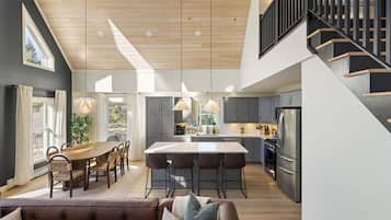 Una cocina moderna con una isla central, un comedor con mesa y sillas de madera, y una escalera con barandilla negra.