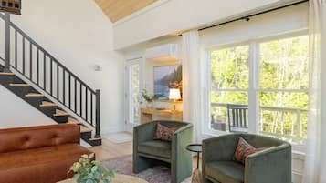 A living room with a brown leather sofa, two green armchairs, a round coffee table, and a view of a forest through large windows.