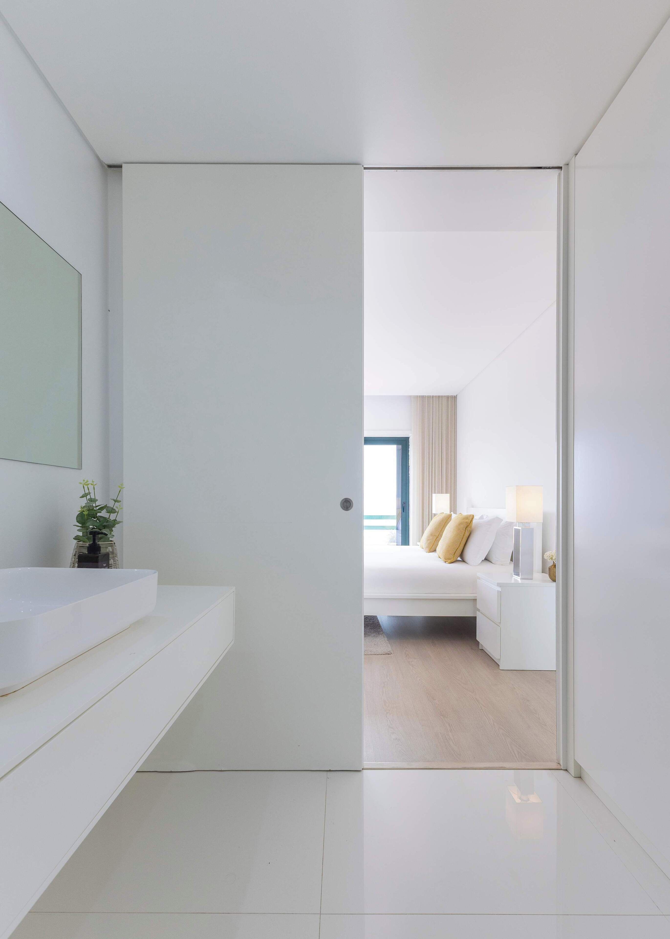 Quarto de hotel moderno com banheiro branco, cama com roupa de cama branca e manta bege, e vista para o mar através de uma janela.