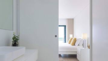 Quarto de hotel moderno com banheiro branco, cama com roupa de cama branca e manta bege, e vista para o mar através de uma janela.