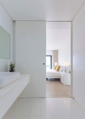 Een moderne hotelkamer met een witte badkamer, een bed met wit bedlinnen en een beige plaid, en uitzicht op de zee door een raam.