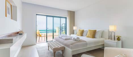 Une chambre d’hôtel moderne avec un grand lit, un coin salon comprenant un fauteuil et une petite table, ainsi qu’un balcon avec vue.