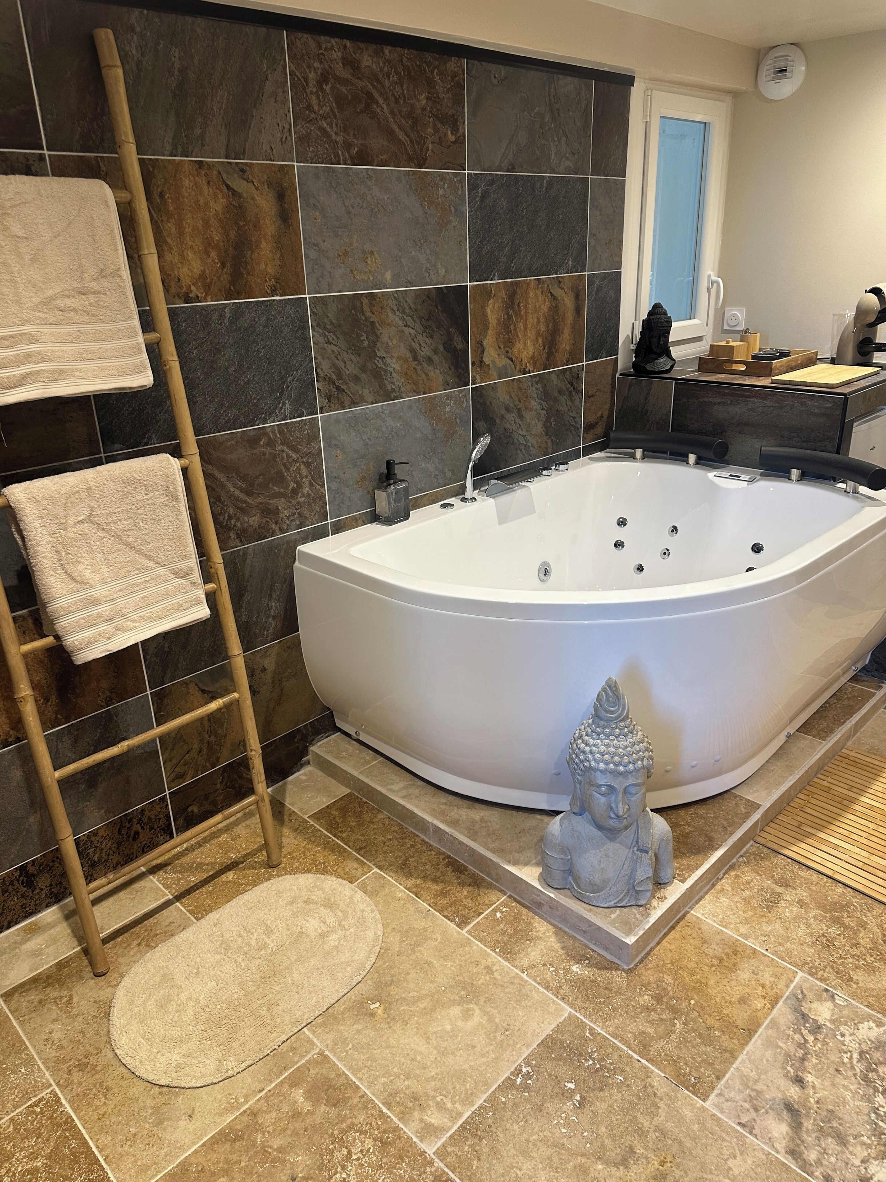 Ein Badezimmer mit einer freistehenden Badewanne, einer Bambusleiter, einer Buddha-Statue und einer Fliesenwand.
