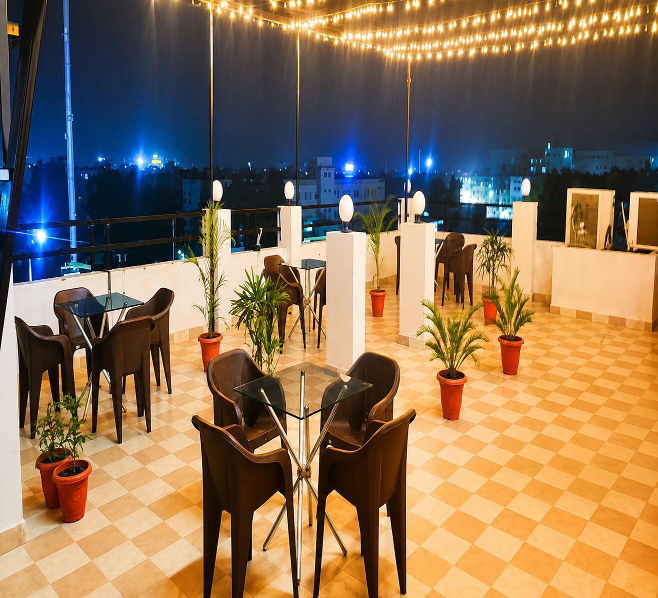Una terraza en la azotea con suelo a cuadros, mesas, sillas, plantas en macetas y luces colgantes.