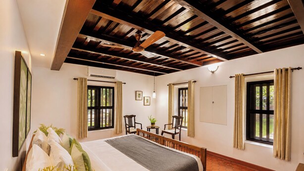 Quarto com cama, duas cadeiras, uma mesinha e um ventilador de teto.